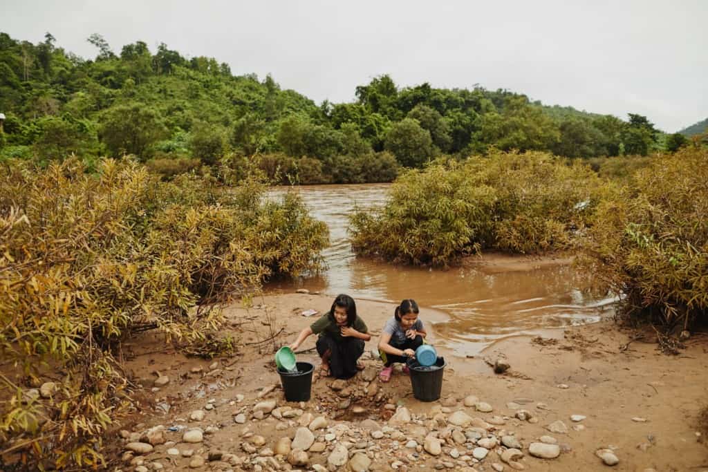 Two adolescent female teen girls, one in a green shirt and black skirt, the other in gray shirt and black pants, sit, squat on the rocky dirt ground holding green and blue pans, pouring water into black buckets. Behind them are a body of brown water, trees and shrubs.