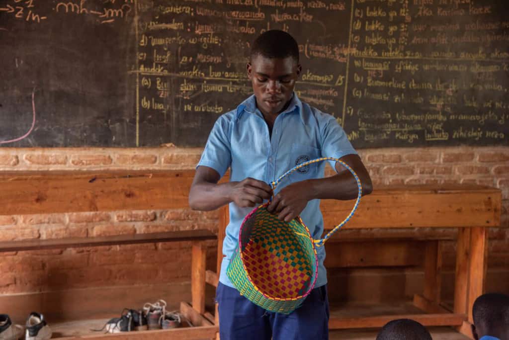 Sebastian is wearing dark blue pants and a blue shirt. He is teaching youth at the Compassion center how to weave bags in one of the classrooms. Behind him is a brick wall with a large black board on it.