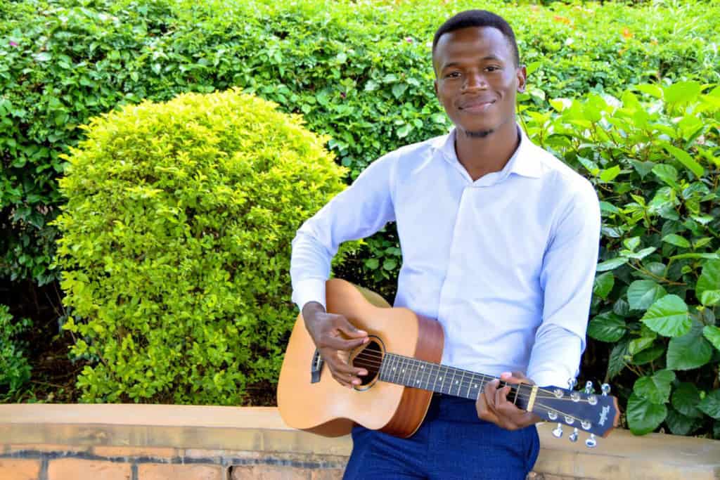 Faida is weairng a white shirt and navy blue pants. He is playing his guitar. Behind him are green bushes.