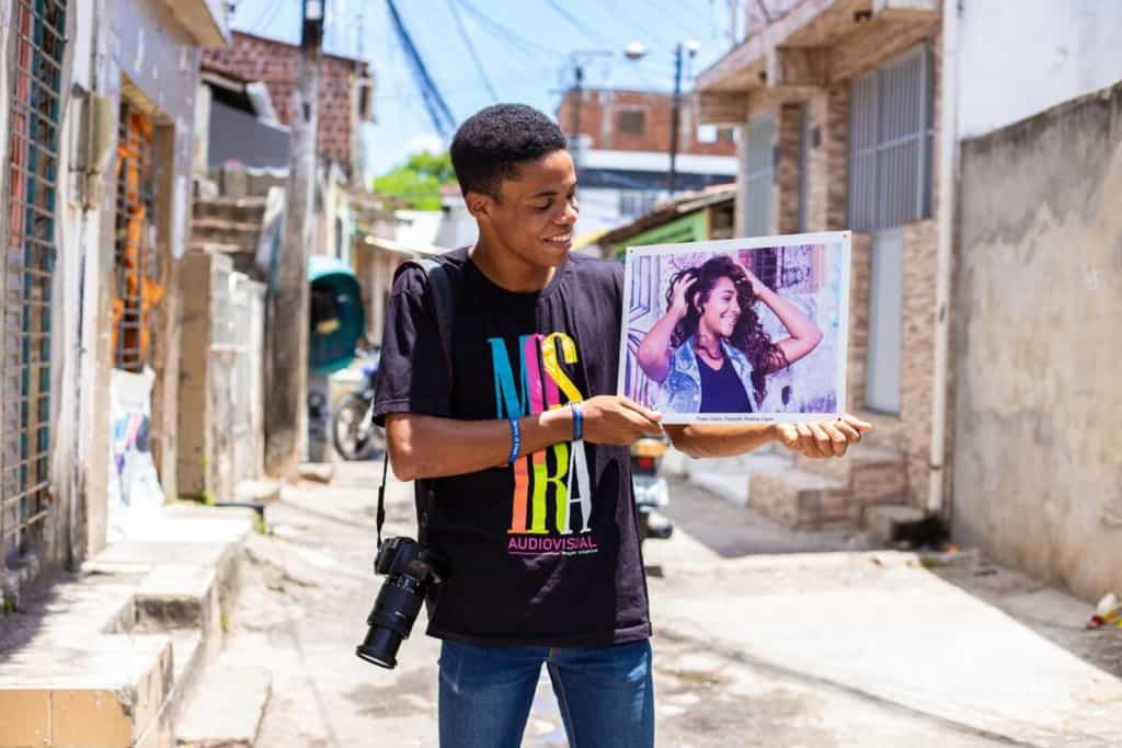 Rhaldney shows one of his favorite pictures. He is wearing a black tee shirt with colored letters, and jeans. He is holding a photo and looking at it.