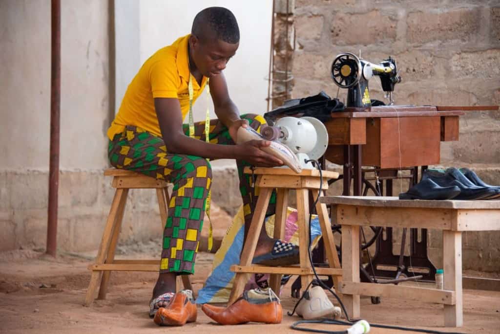 Theophile is wearing a yellow shirt. He is using a machine to polish a pair of shoes he is making.