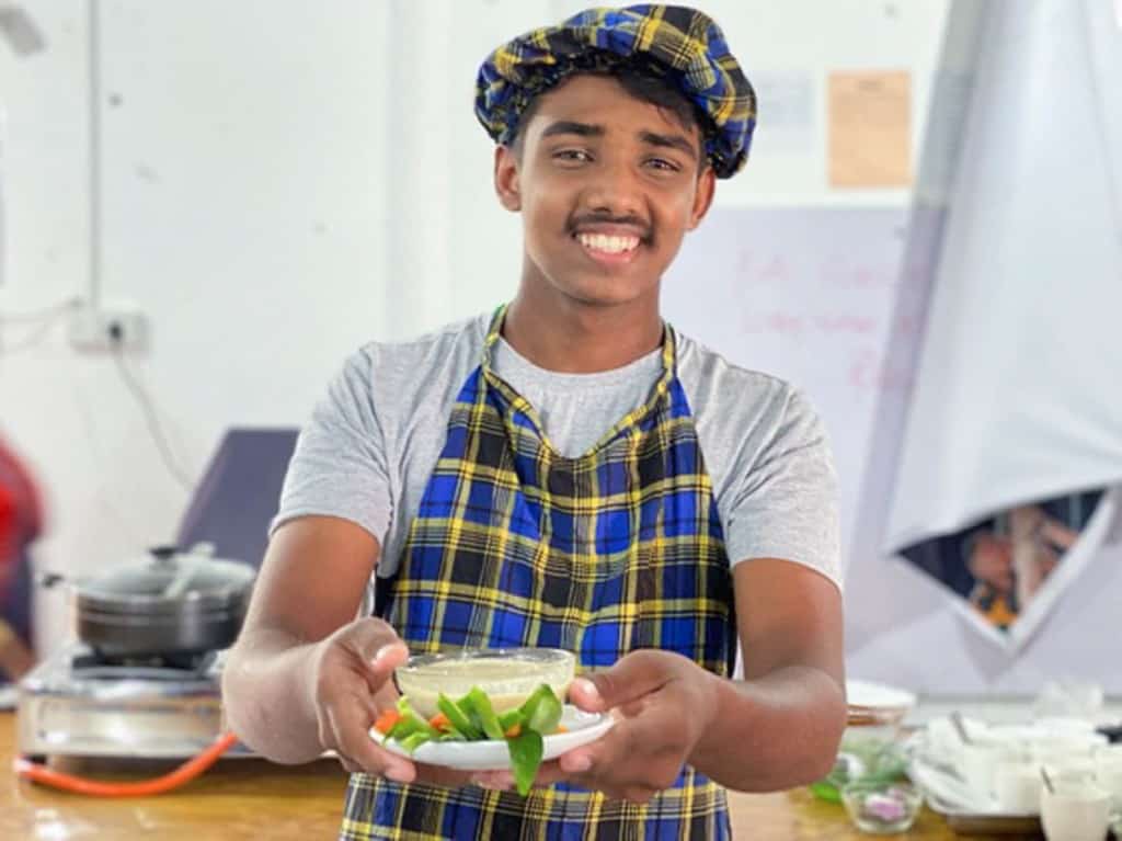 Piyeston is wearing a gray shirt with blue and yellow plaid apron. He is holding out a bowl of soup that he made.