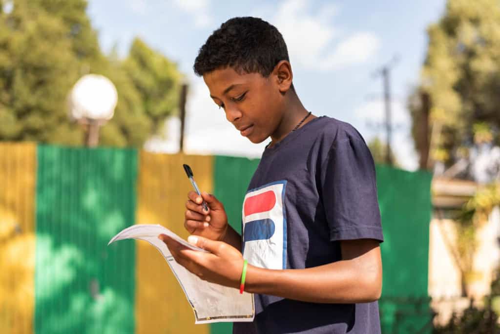 Young man wearing a blue shirt. He is standing outside and is writing a letter to his sponsor.