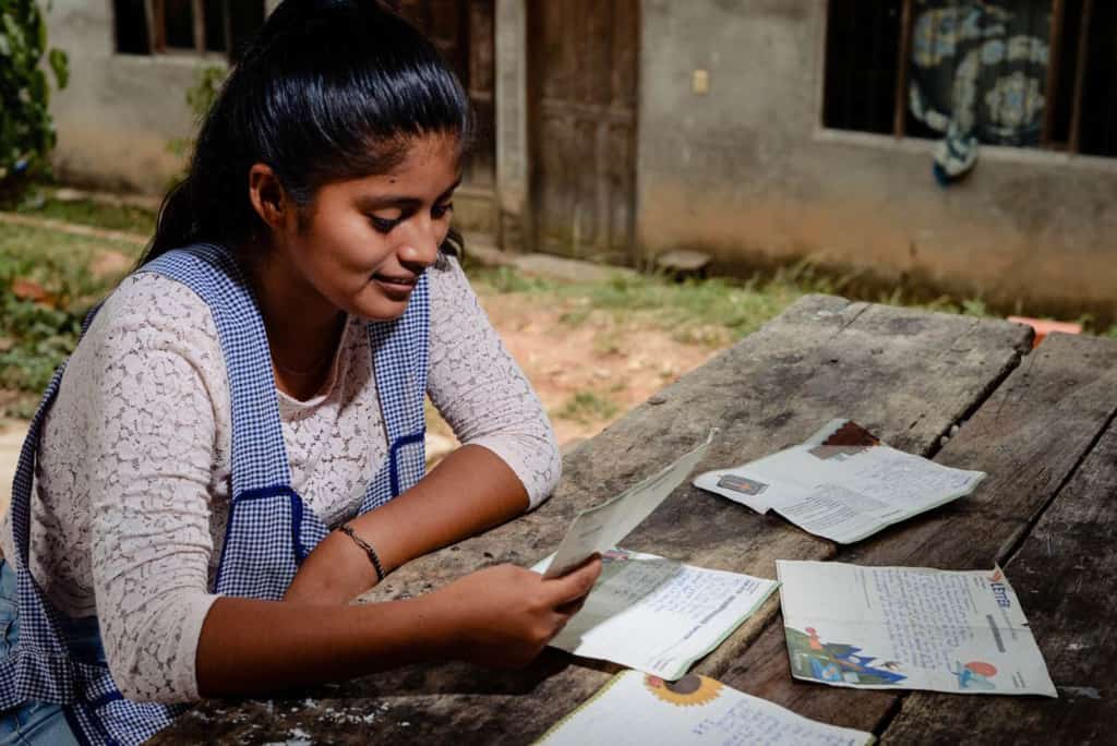 Young woman wearing a white shirt and a blue and white checkered apron. She is sitting at a table outside and is reading letters from her sponsor.