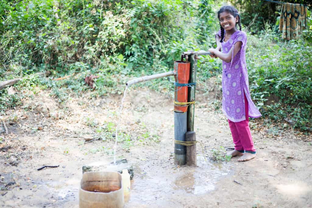 Girl smiling at the camera as she pumps water into a wooden bucket that is on the ground. She is wearing pink pants and a purple dress. There are trees in the background.