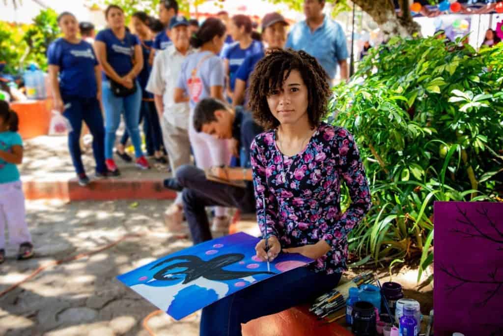 Sandra is wearing a black dress with pink and blue flowers on it. She is painting at the craft fair and she is sitting in front of a tree. There are many other people in the background.