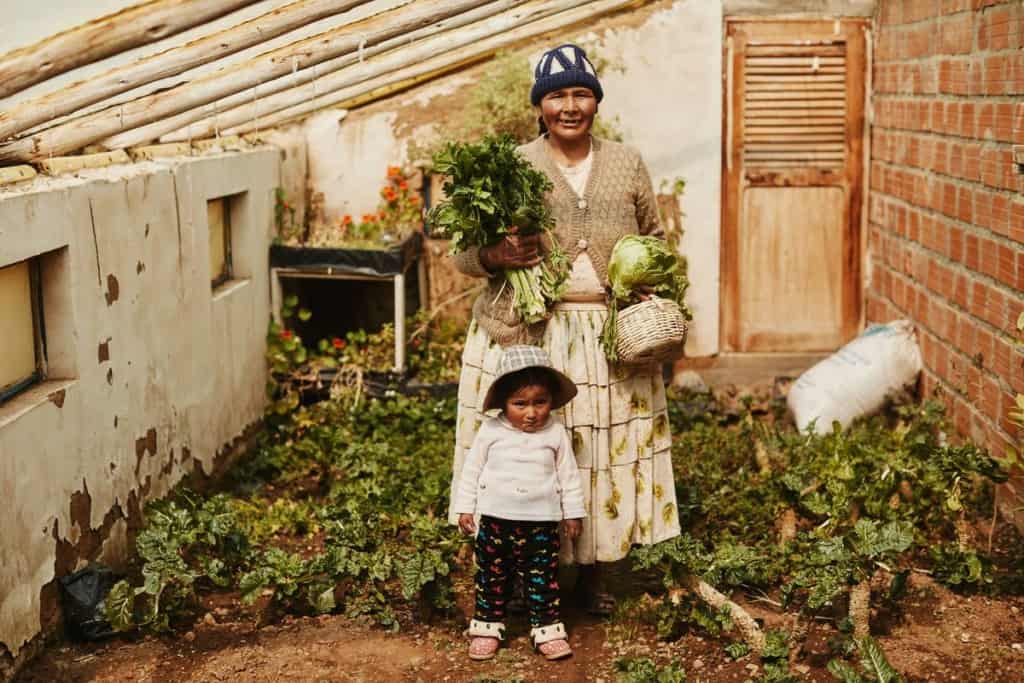 A woman wearing a long skirt, sweater and hat holds vegetables in a garden in Brazil. A toddler wearing a hat stands near her.