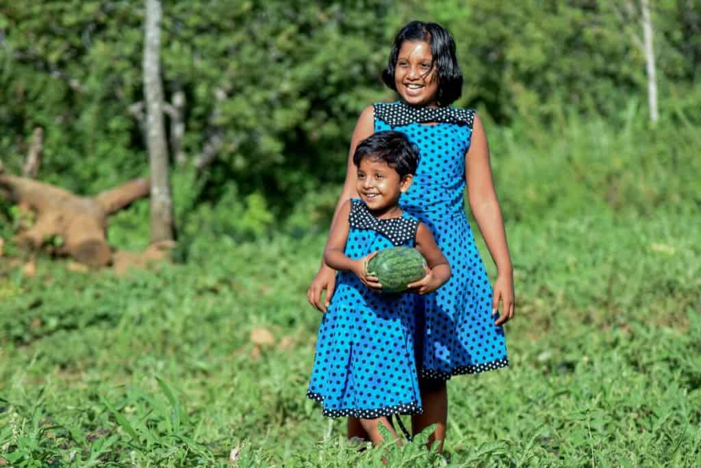 Two sisters wearing matching blue and black polka dot dresses are in a garden in Sri Lanka. The younger one is carrying a watermelon she picked.