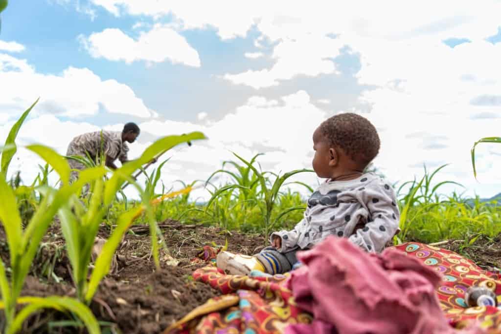 A baby sits on a blanket spread out in a garden in Tanzania as her parent works with a hoe nearby