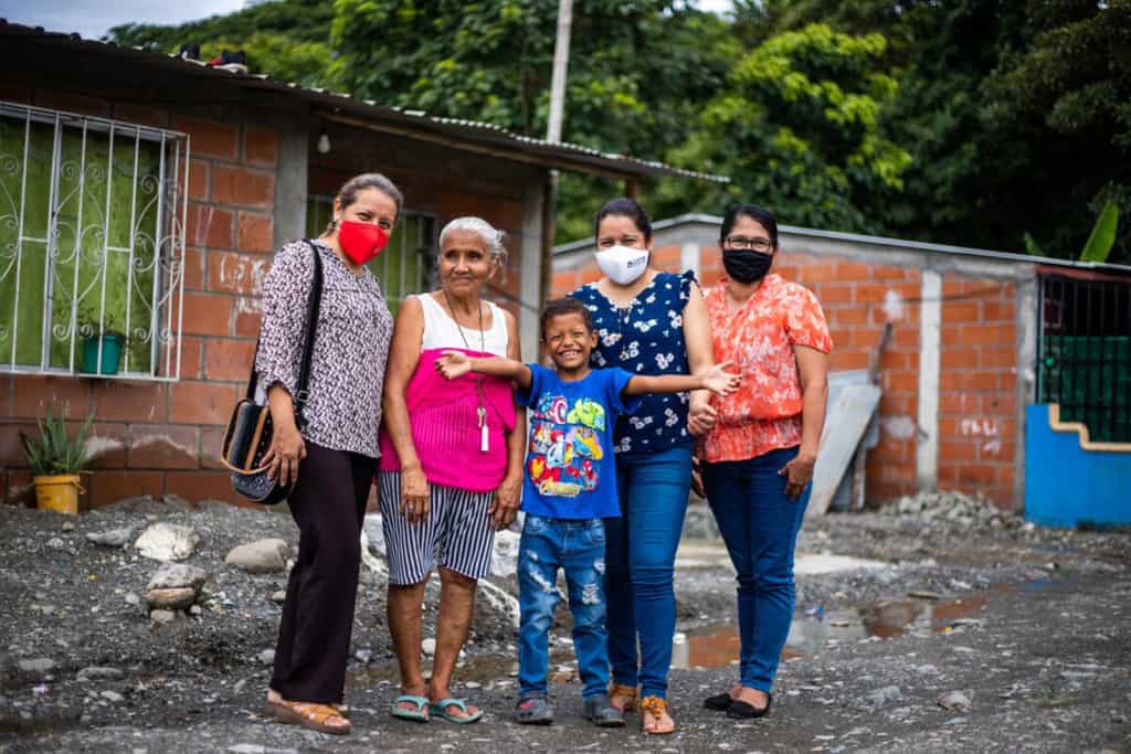 Jesus, wearing a dark blue shirt and jeans, is standing with his grandmother, Rosa, wearing a pink and white shirt and black and white striped shorts. Next to them is the project director and the tutors. They are standing outside Jesus' home. The Compassion staff are wearing face masks.