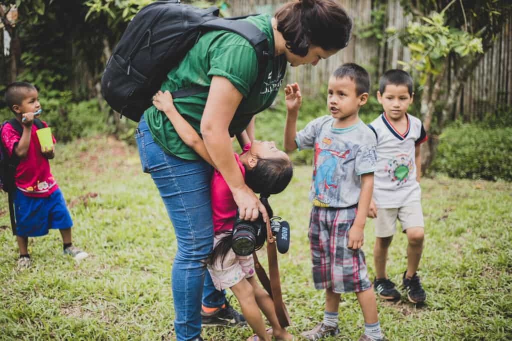 Sara Navarro, Compassion photojournalist leans over to hug a child while holding her camera and wearing a backpack. Other children walk in the grass on the background.