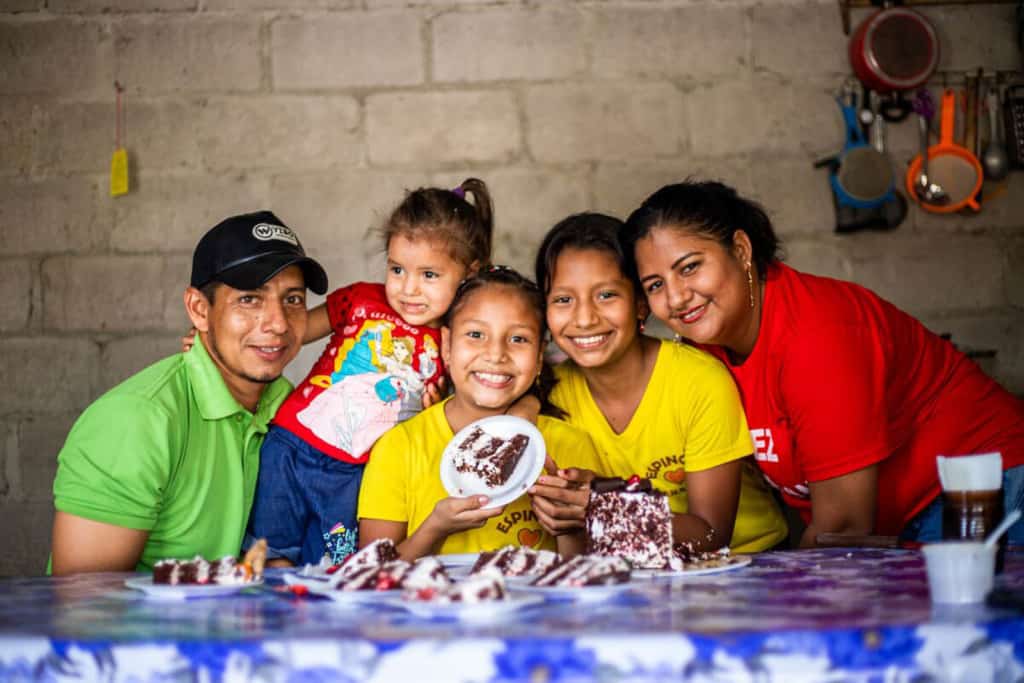 Ashley and Emily are wearing yellow shirts and jeans. They are surrounded by their younger sister and parents. They are all sitting at a table in their kitchen. Ashley is holding up a piece of her birthday cake.