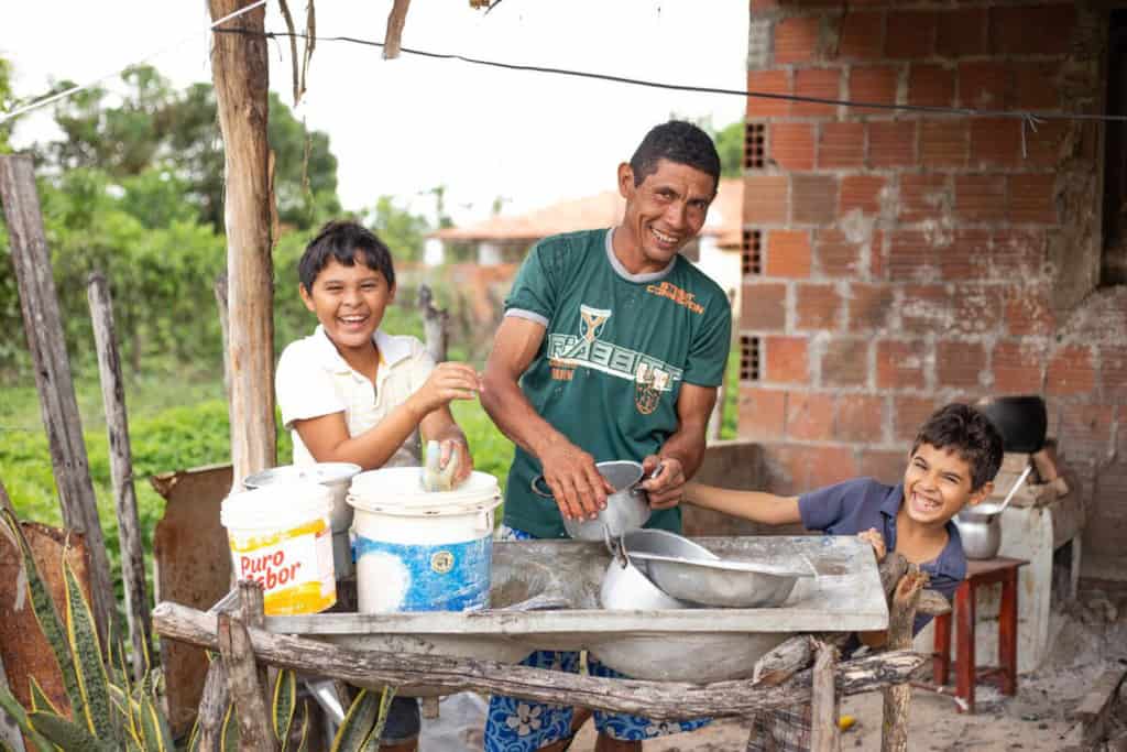 Damião, wearing a green shirt, is with his sons, Davi, wearing a white shirt, and Luan, wearing a blue shirt. They are washing dishes together in their back yard.