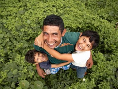 Damião, wearing a green shirt, is with his sons, Davi, wearing a white shirt, and Luan, wearing a blue shirt. They are hugging each other in their back yard. They are standing in the grass.