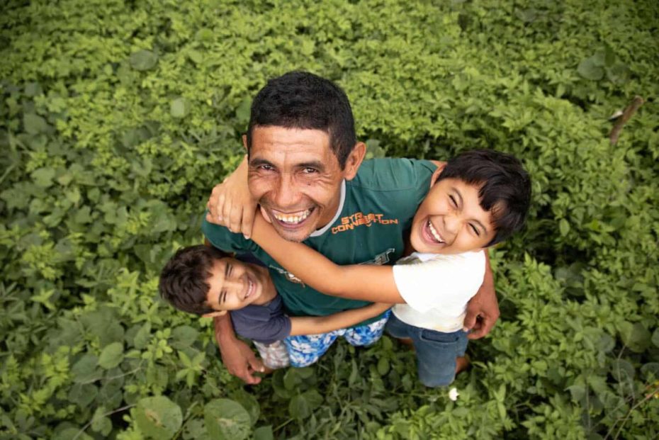 Damião, wearing a green shirt, is with his sons, Davi, wearing a white shirt, and Luan, wearing a blue shirt. They are hugging each other in their back yard. They are standing in the grass.