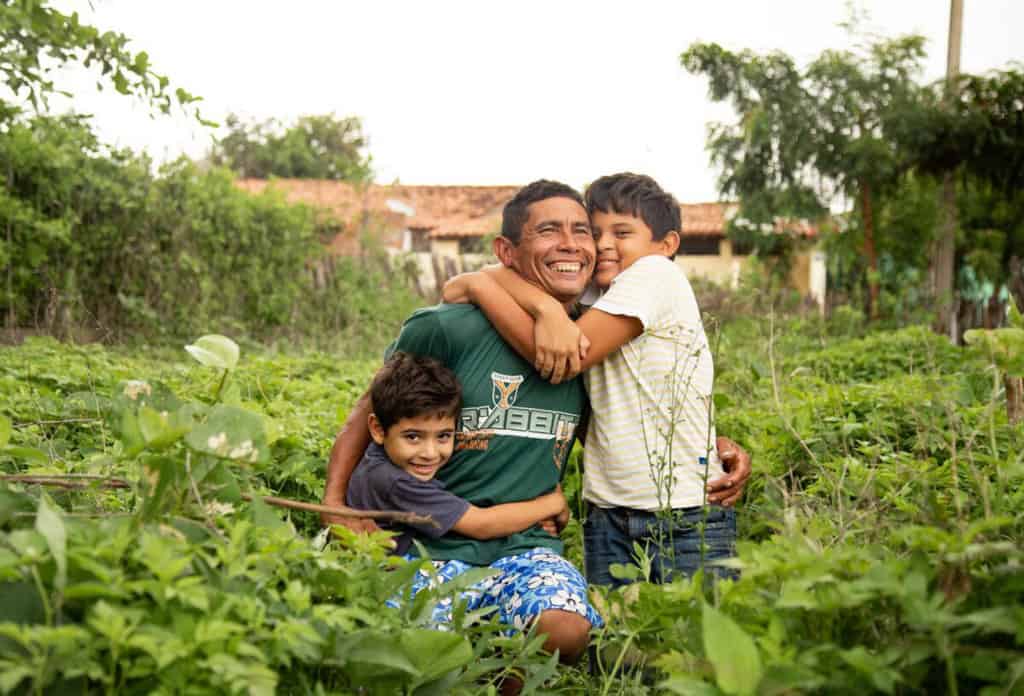 Damião, wearing a green shirt, is with his sons, Davi, wearing a white shirt, and Luan, wearing a blue shirt. They are kneeling down in the grass in their backyard and are hugging each other.