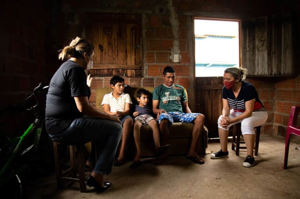 Damião, wearing a green shirt, is with his sons, Davi, wearing a white shirt, and Luan, wearing a blue shirt. They are sitting inside their home with two Compassion volunteers who are praying for them. The volunteers are wearing face masks.