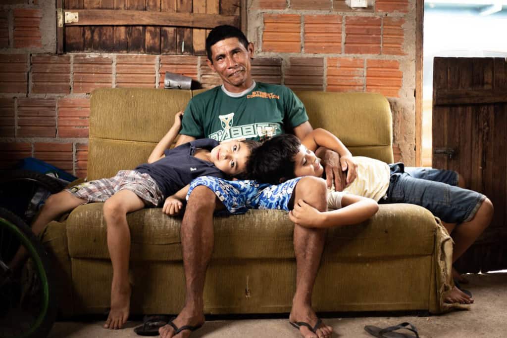Damião, wearing a green shirt, is with his sons, Davi, wearing a white shirt, and Luan, wearing a blue shirt. They are sitting together on a couch in their home and Davi and Luan are resting their heads in their dad's lap.