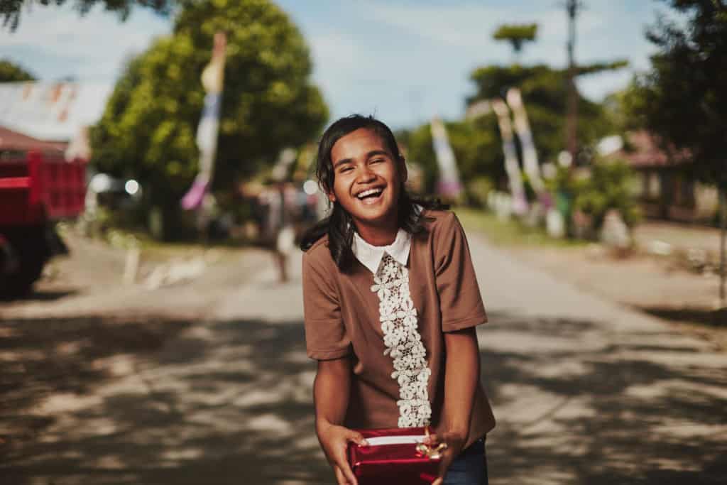 A laughing girl child wearing a brown shirt with white lace and collar stands outside in the middle of a street holding a present wrapped in shiny red wrapping paper. Behind her is blue sky and trees.