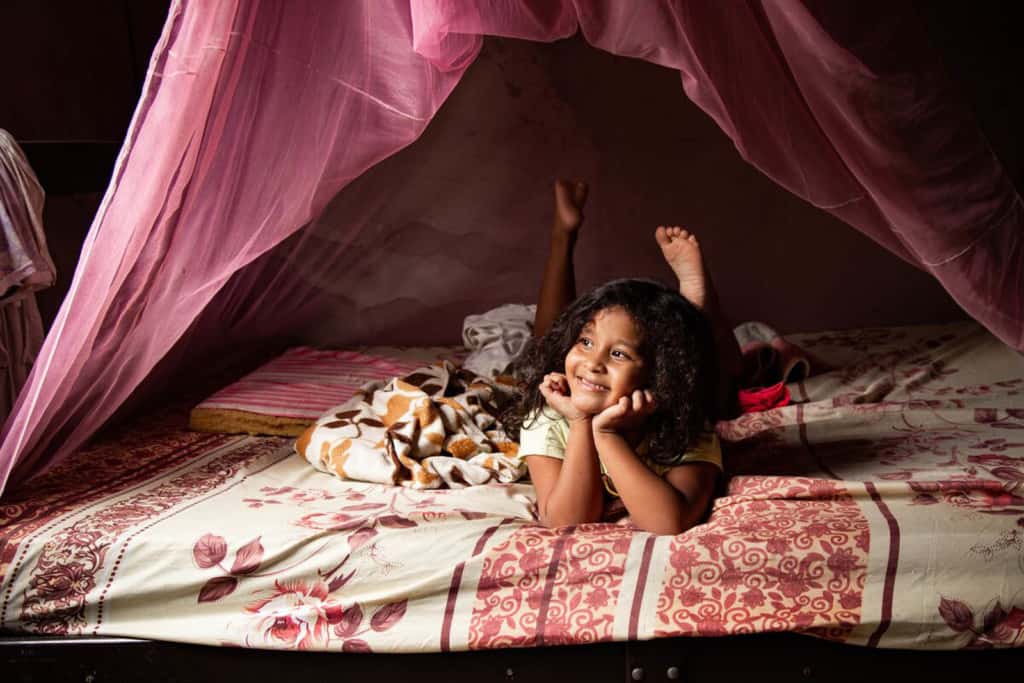 Sophia is wearing a cream colored shirt and shorts. She is laying on her bed and there is a pink mosquito net above her.