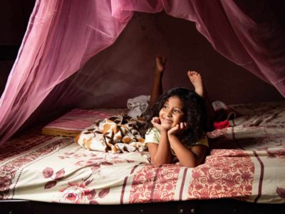 Sophia is wearing a cream colored shirt and shorts. She is laying on her bed and there is a pink mosquito net above her.