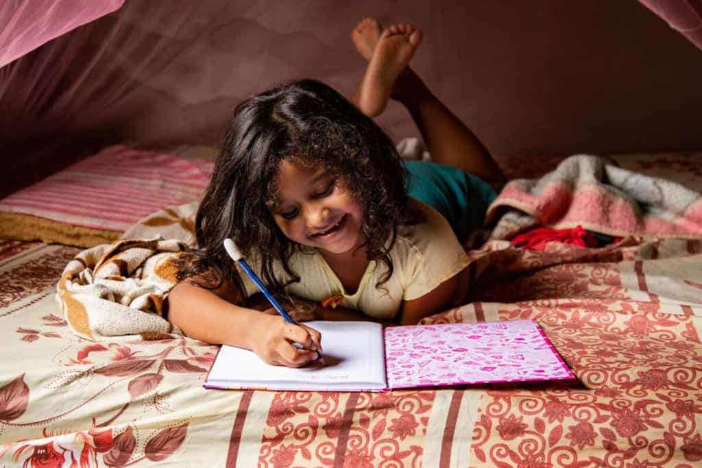 Sophia is wearing a cream colored shirt and shorts. She is laying on her bed and there is a pink mosquito net above her. She is writing in a notebook.