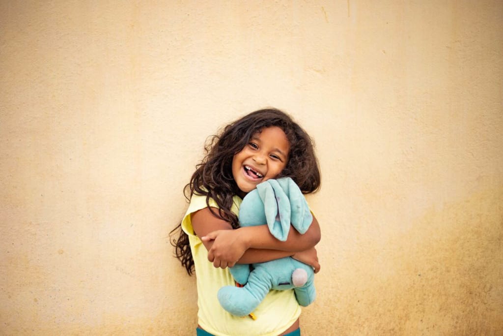 Sophia is wearing a yellow shirt. She is standing in front of a tan wall and is hugging her favorite stuffed animal.