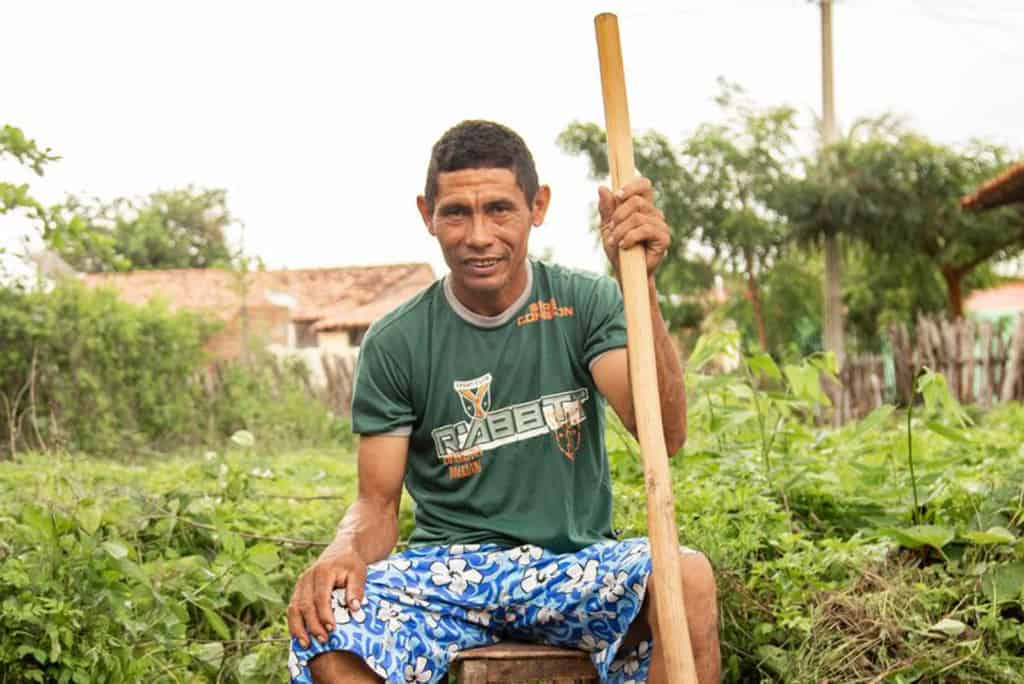 Damião, wearing a green shirt and blue patterned shorts, is sitting on a stool outside in his back yard. He is holding a large stick