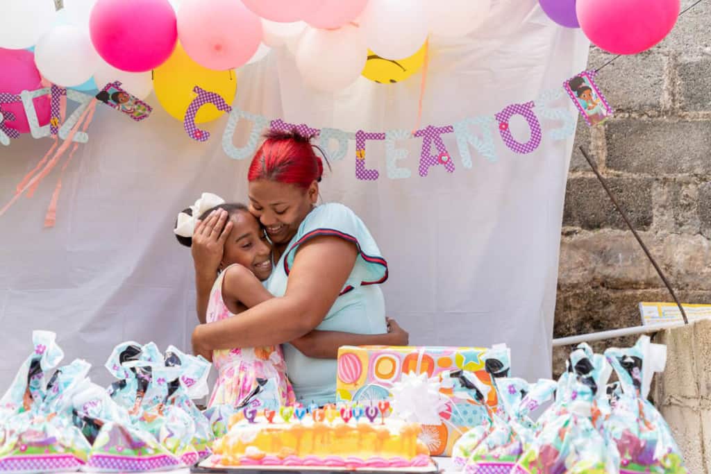 Lorielky is wearig a white dress with pink and orange flowers. She is standing behind a table with a birthday cake and gifts on it. Abover her are colorful balloons and behind her is a sign that says, "Cumpleanos." She is hugging her mother, Delmi.