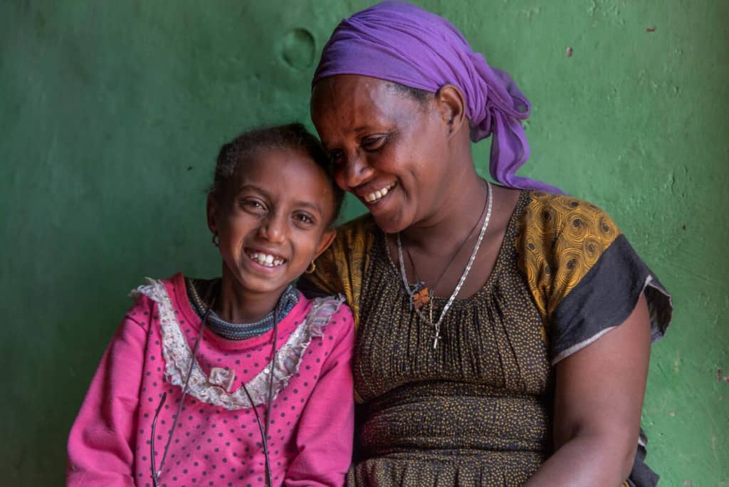 Helen is wearing a yellow and brown dress and a purple head covering. She is sitting next to her daughter, Rediet, wearing a pink shirt. The background is a green wall.