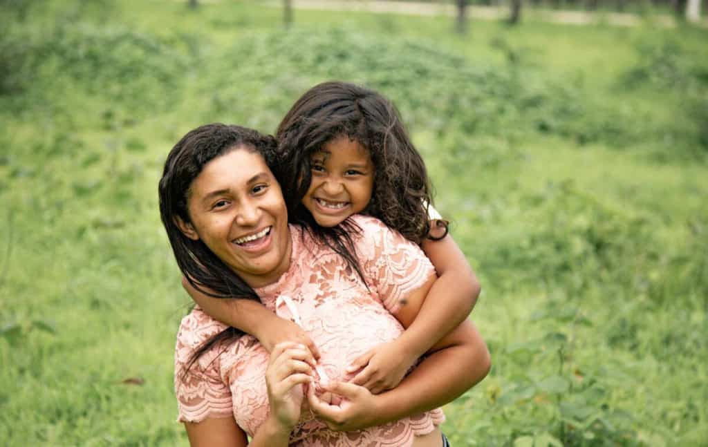 Spohia is standing outside her home in the grass with her mother, Milena. Milena is wearing a pink lace shirt. Sophia is standing behind Milena with her arms around her.