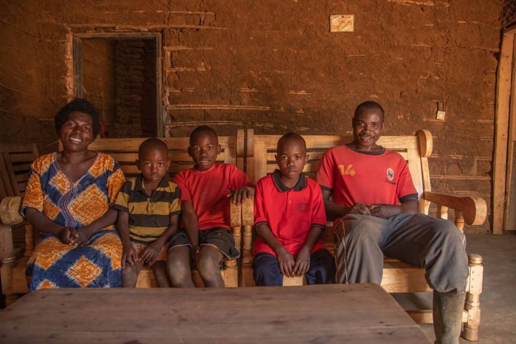 Frank is wearing a red shirt and dark shorts. He is sitting on a bench with his family in front of their home, which has been renovated using money from a gift from Frank's sponsor.