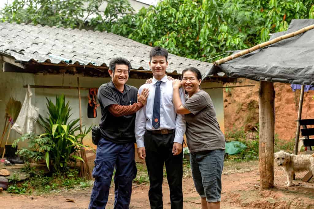 A family portrait of Surayut wearing his school uniform with his parent with smile, standing in front of his house.