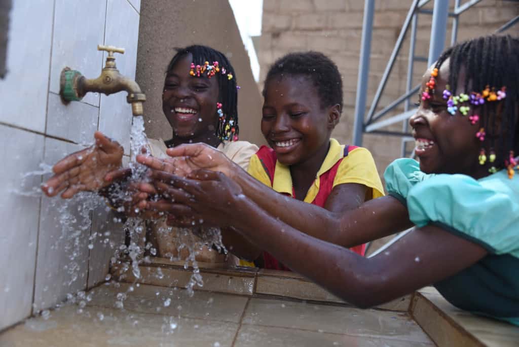 A group of girls children students one is wearing a red and yellow shirt another a green dress with her hair in colorful braids looks at the running flowing water smiling as they are splashing washing hands at a station with clean running water from a faucet at a sink facility at the center practicing good personal hygiene washing and cleaning over a new outdoor outside sink tiled area.