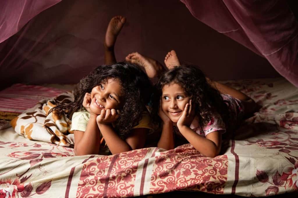 Sophia is wearing a cream colored shirt and shorts. She is laying on her bed and there is a pink mosquito net above her. Next to her is her sister, Daphyne. Both of them are resting their hands on their chins.