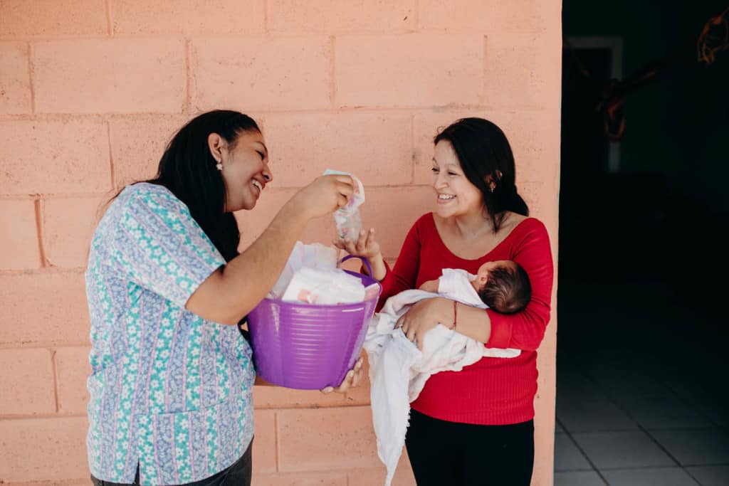 A Compassion staff member is giving Brenda, in a red shirt, a gift for baby Samantha Isabel, who she is holding in her arms.