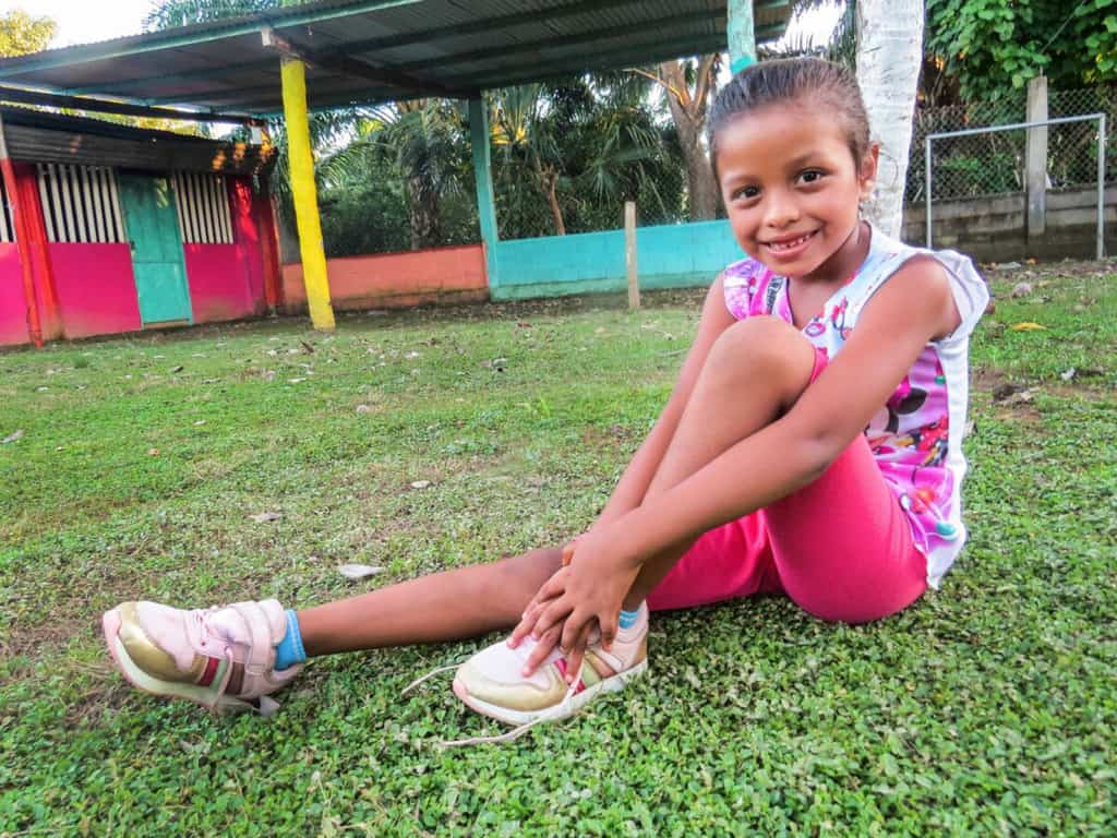 Iris is wearing a brightly colored shirt and pink shorts. She is sitting in the yard of the Compassion center and is showing off her new shoes.