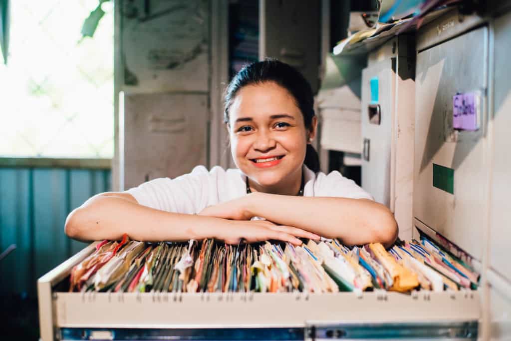 Sandy, wearing a white shirt, stands smiling in an office with arms on an open file cabinet drawer filled with files and paperwork.