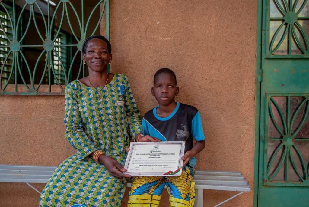 A strong mother wearing a green, blue and white dress smiles proudly as she and her son hold up a certificate of recognition of academic excellence from Burkina Faso's president.