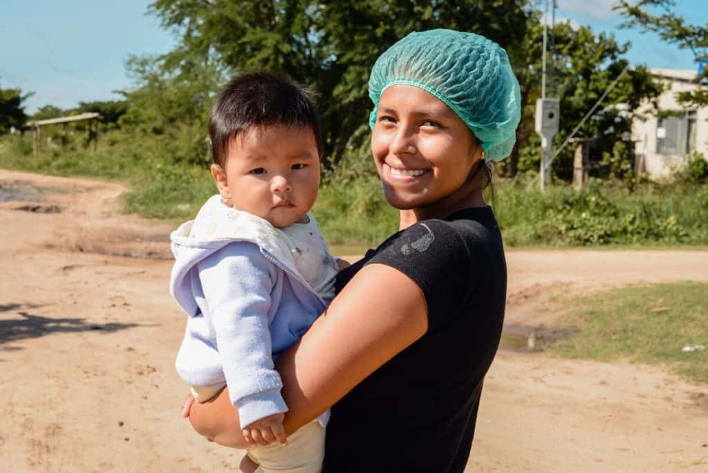 A teen mom in Bolivia wearing a green hair net and black shirt holds her young child.