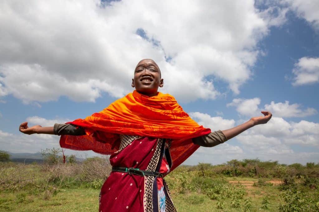 A Kenyan boy in orange and red clothing holds his hands out and smiles. His eyes are closed and he is gazing upward