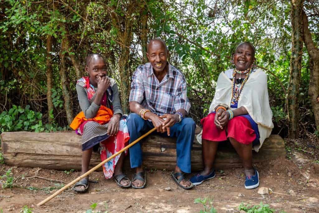 A teenage boy and his two parents sit on a log outside. They are all smiling.