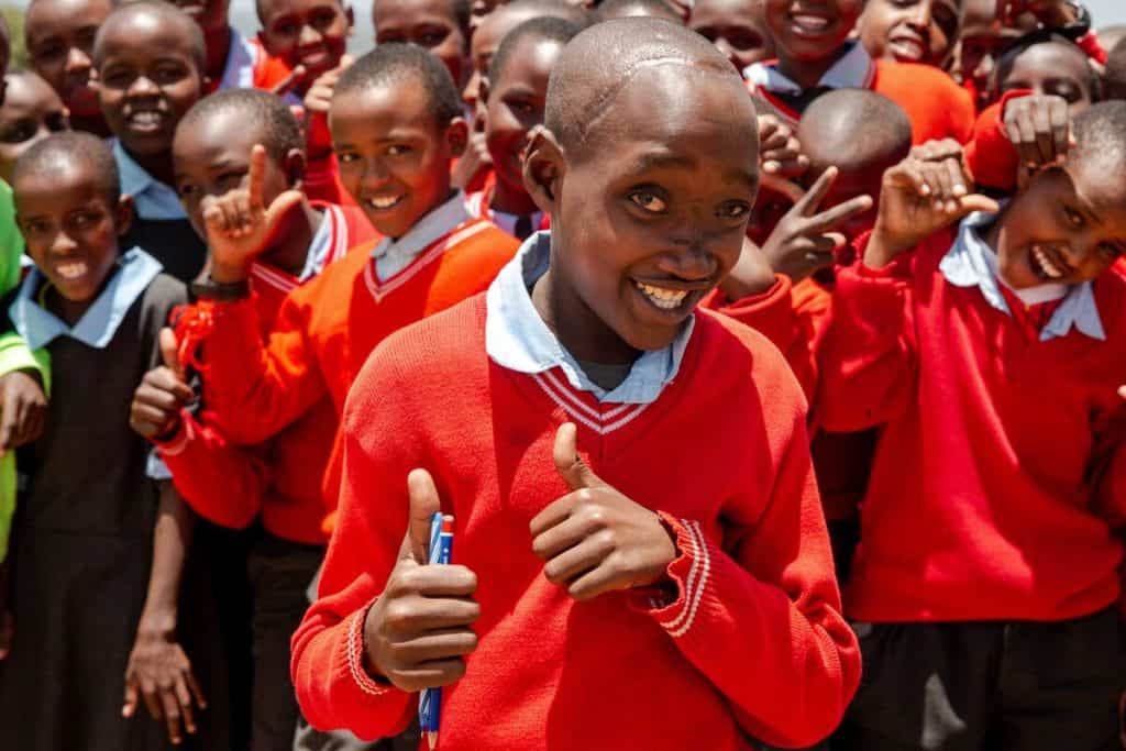 A Kenyan teenager gives two thumbs up. A group of children stands smiling behind him. They are wearing red sweaters over white collared shirts. 