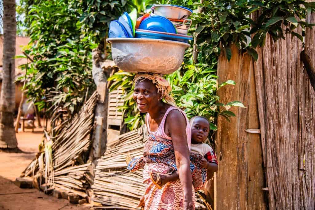 A woman walks with a toddler strapped to her back in a colorful cloth. She is balancing a large pan stacked high with dishes on her head.