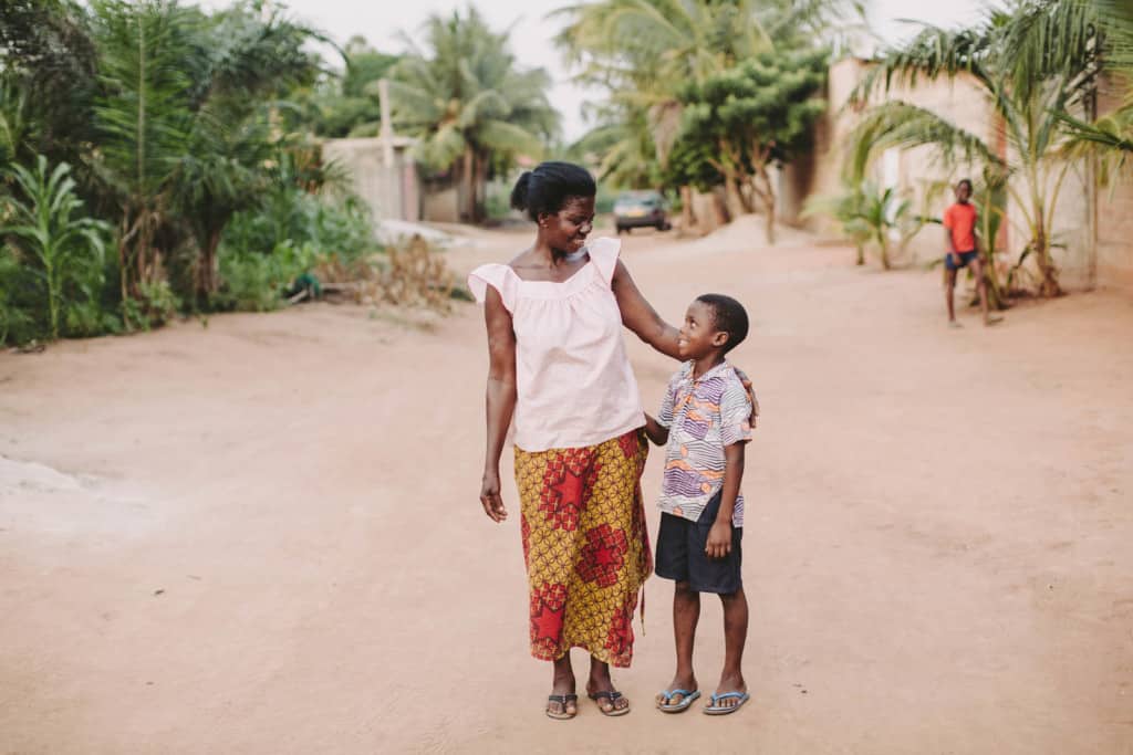 Boy wearing a blue and orange pattern shirt smiles standing beside his mother.