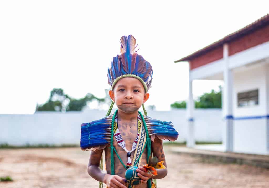 Kaio is standing outside the Compassion center and is wearing a traditional Guajajara costume. He is also holding a rattle. His face and body are painted with designs.