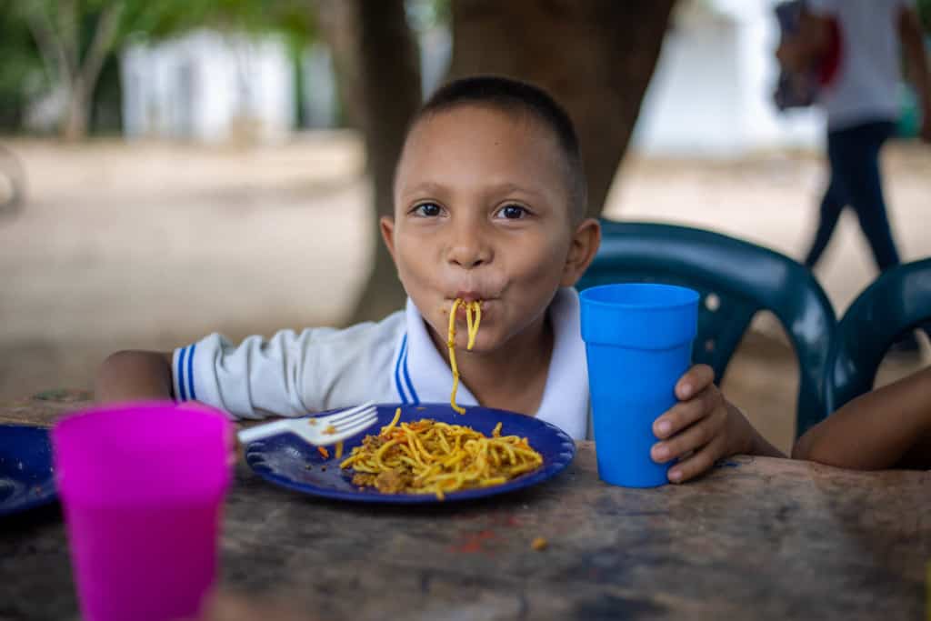 Marcos, wearing a white shirt, is eating spaghetti. He is sitting at a table and is holding a blue cup.