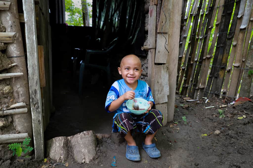 Axel is wearing a light blue and white soccer jersey and black shorts. He is sitting in his house’s doorway eating red beans with sour cream, which is one of his favorite foods. There is a fence beside the home.