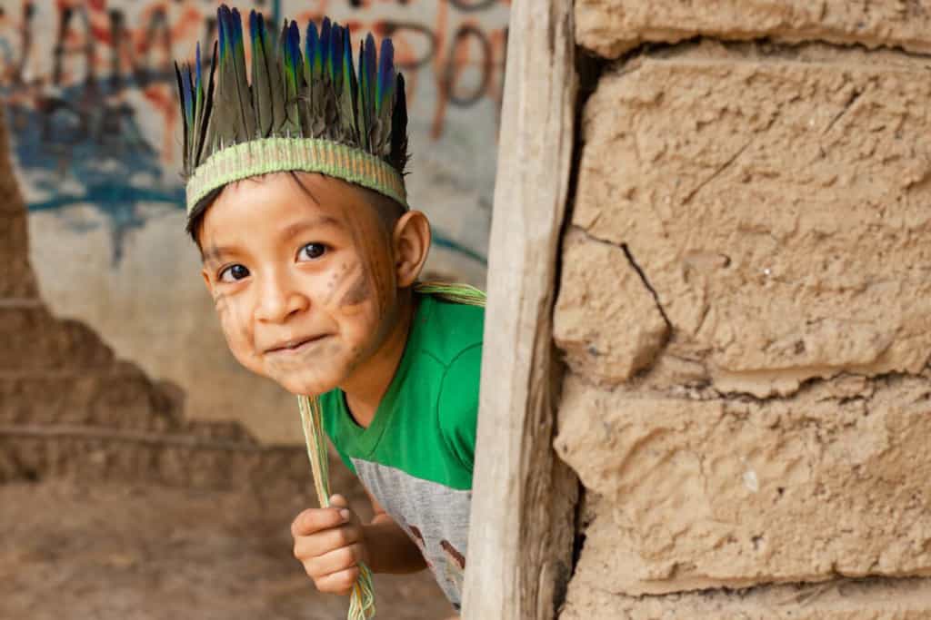 Kaio is looking around the corner outside his home. He is wearing a green and gray shirt as well as a traditional head covering. His face is also painted.