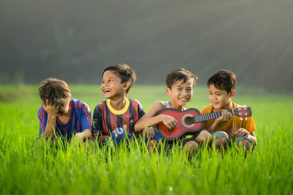 Four boys sitting in a green field. One is playing a small stringed instrument while the others smile and laugh.